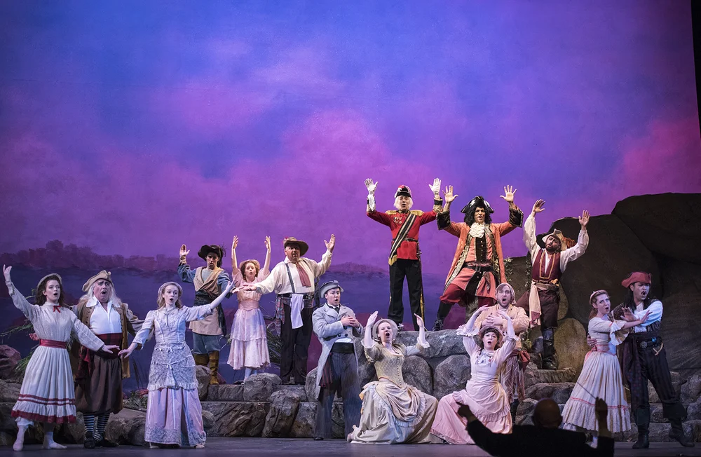 A large group of actors in period costumes on stage with raised hands, performing a chorus against a purple sunset backdrop.