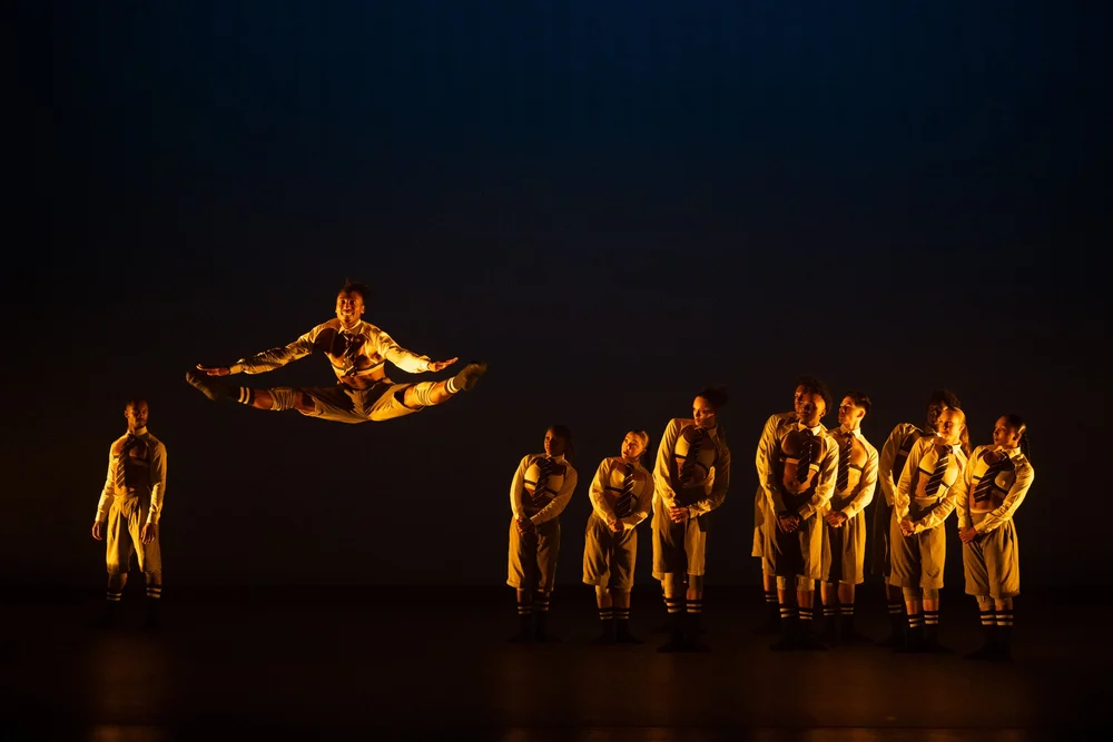 Dancer in a gold costume leaps mid-air over a line of fellow performers on a dark stage.