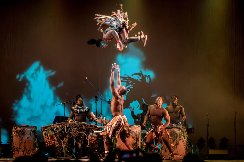 A shirtless dancer leaps overhead as another performer soars midair during a stage drum performance with dancers in foliage costumes on a dimly lit set.