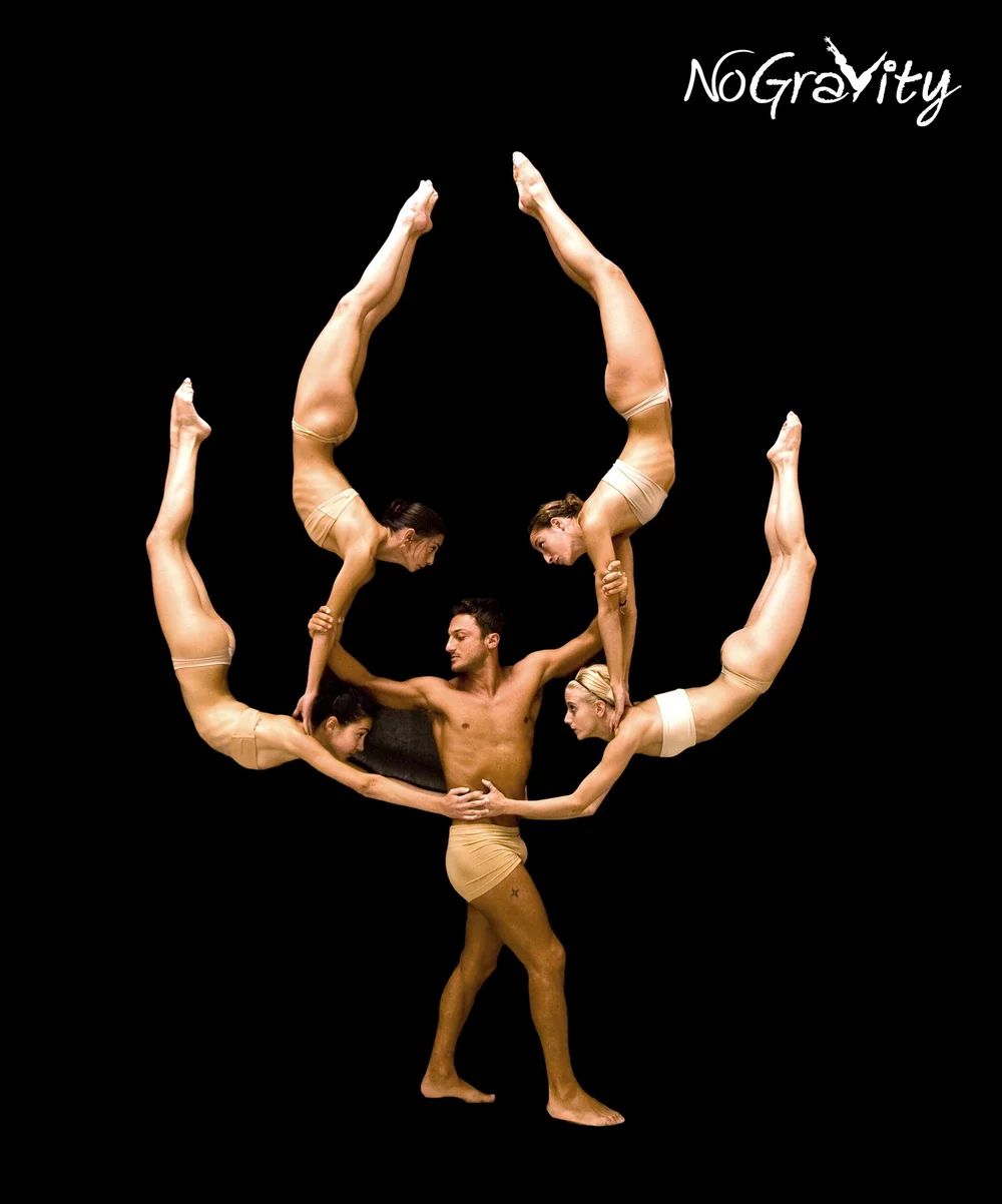 Five nude-toned acrobats form a circular human arch around a male base on a black background, performing a synchronized balance, with No Gravity logo in the corner.