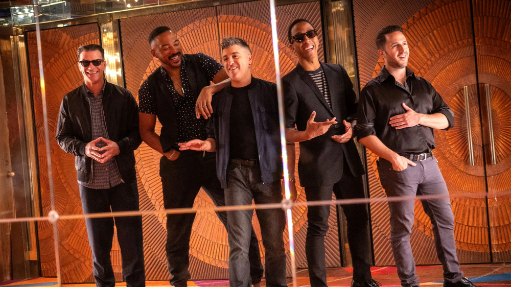 Five men stand together in a warmly lit lobby with circular orange wall panels, posing and smiling.