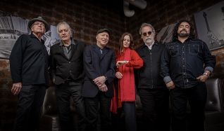 Seven-member band lineup posing in a dim, brick-walled venue; six men and one woman, the woman in red, all in dark clothing.