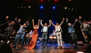 Diverse stage ensemble of singers and musicians standing in a line with arms raised in celebration after a live performance on a lit theater stage.