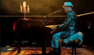 A man in a blue hat and patterned shirt plays a grand piano on a dimly lit stage, candlelight illuminating the scene.