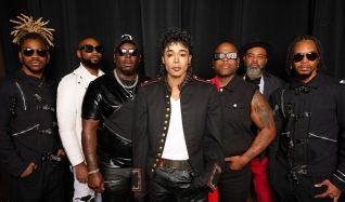 Promo photo of a nine-member music group posing against a black backdrop, with a woman center-front wearing a white top and leather jacket.